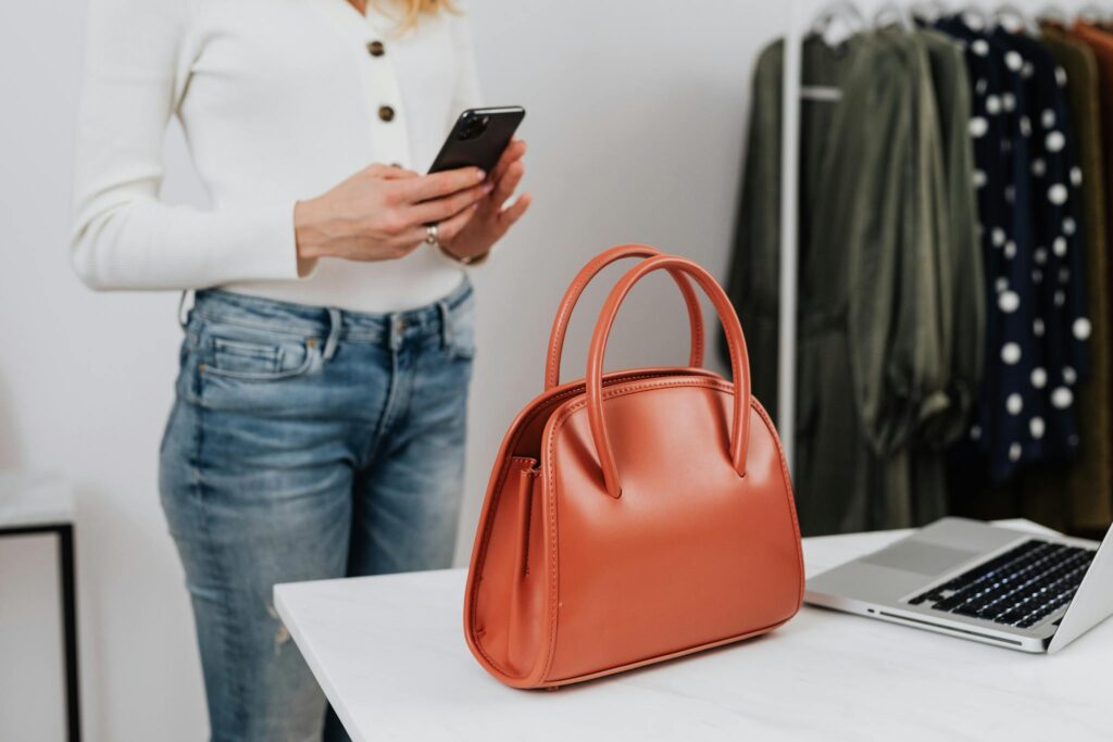 Woman in white long sleeves using smartphone in fashion studio with handbag on table.